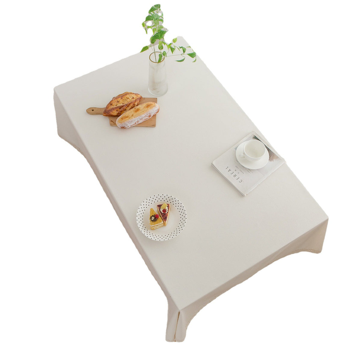 A rectangular beige tablecloth draped over a table, styled with a small vase of green leaves, a sandwich on a wooden board, a small plate, and a cup and saucer, set against a plain white background.