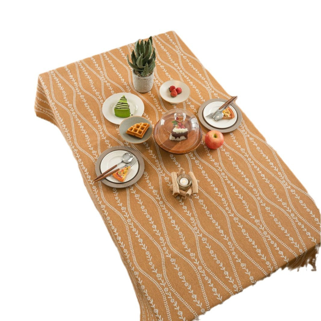 A tan woven table runner with a white geometric leaf pattern, styled with miniature plates, a small plant, and dessert props on a white background.
