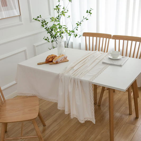 A cream-colored tablecloth with delicate lace trim draped over a white dining table with wooden chairs in a bright, modern room.