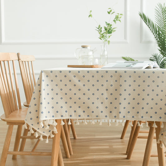 A cream-colored tablecloth, adorned with small blue daisies and fringed borders, is placed beside a wooden dining table, complementing the modern and bright room with the matching wooden dining chairs.
