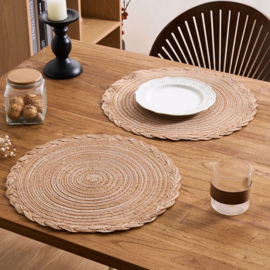 Two round woven natural fiber placemats on a wooden dining table, styled with a white plate on one placemat and a glass cup nearby.