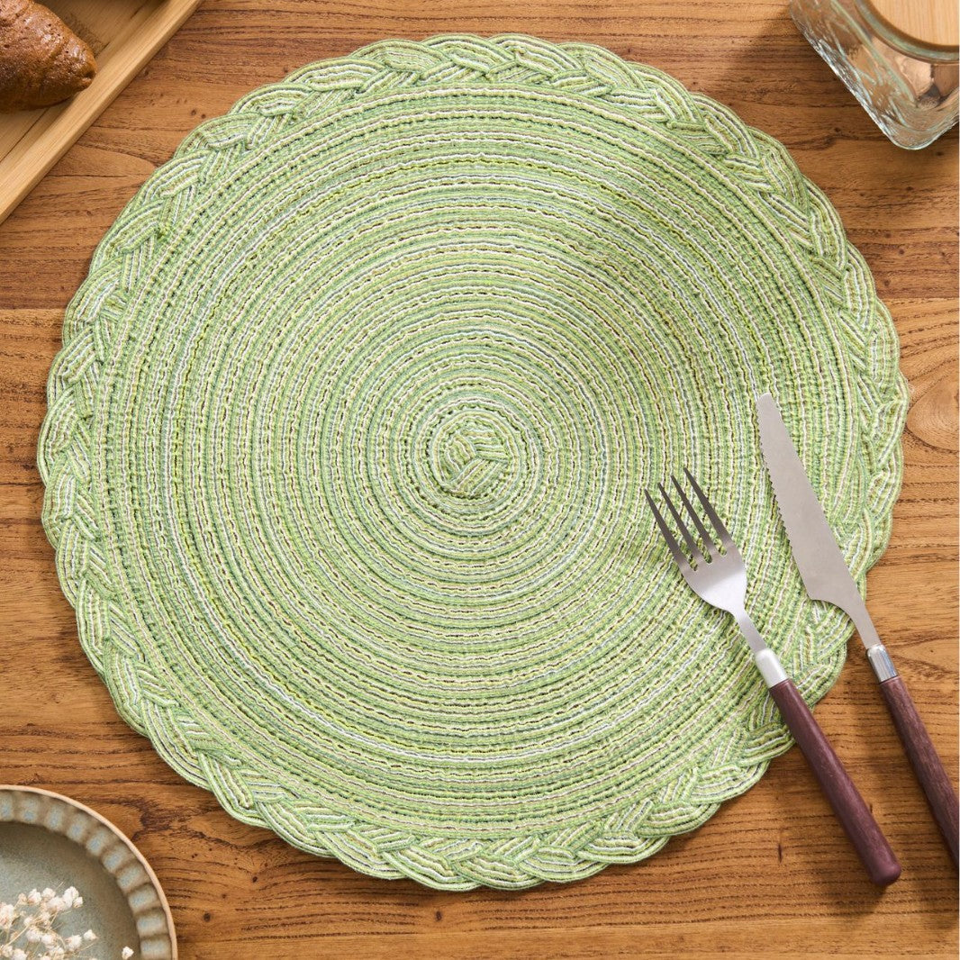 A round woven placemat in light green with braided concentric patterns and scalloped edges, styled on a wooden table with a fork and knife on top.
