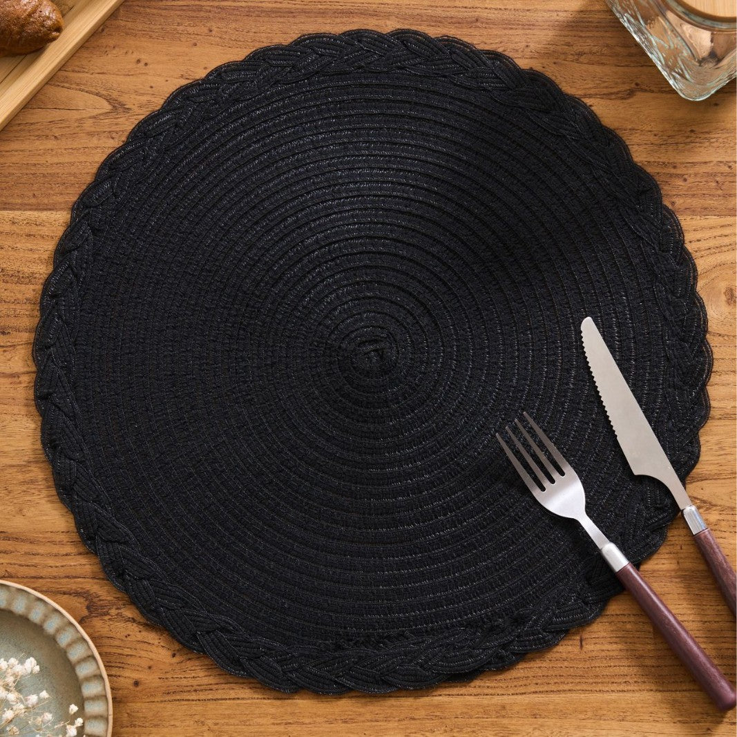 A round black braided placemat on a wooden table, styled with a fork and knife that have purple handles.