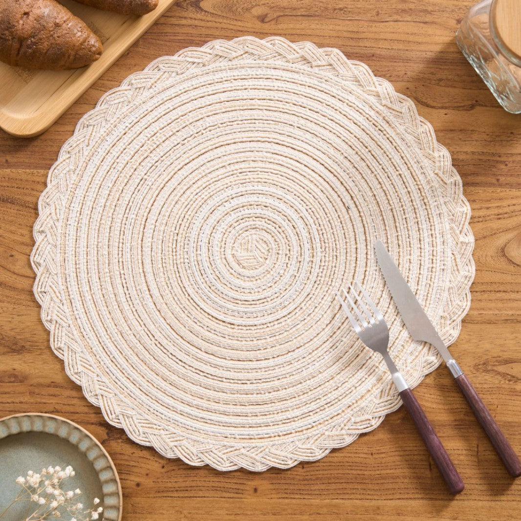 Round woven placemat on a wooden table with cutlery and bread.