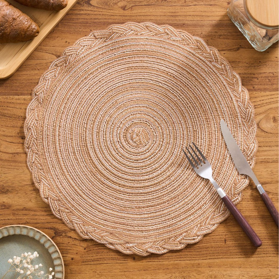 A round woven placemat in a natural tan jute style with braided edges, displayed on a wooden table with a fork and knife on top.