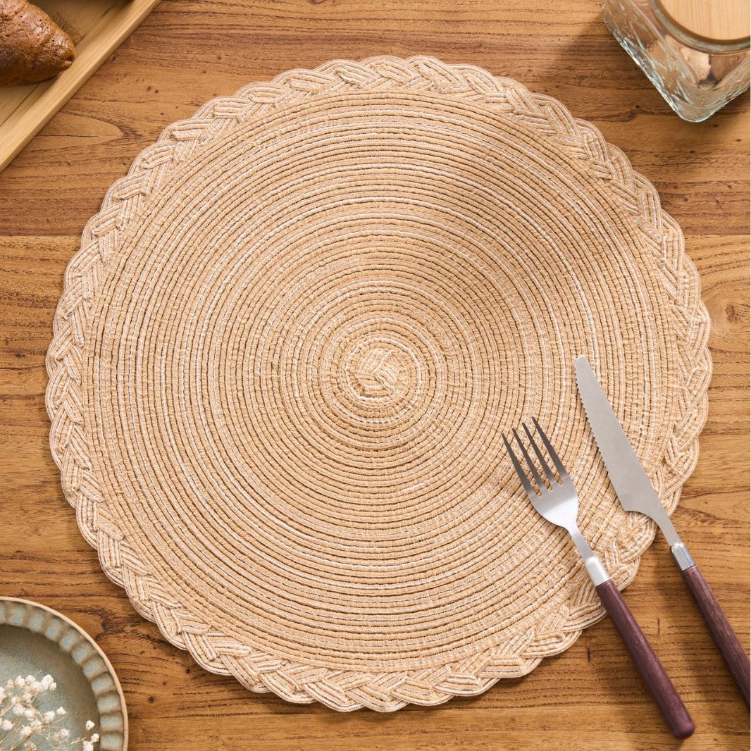 A round woven placemat in a natural beige straw color, styled on a wooden table with a fork and knife on top.